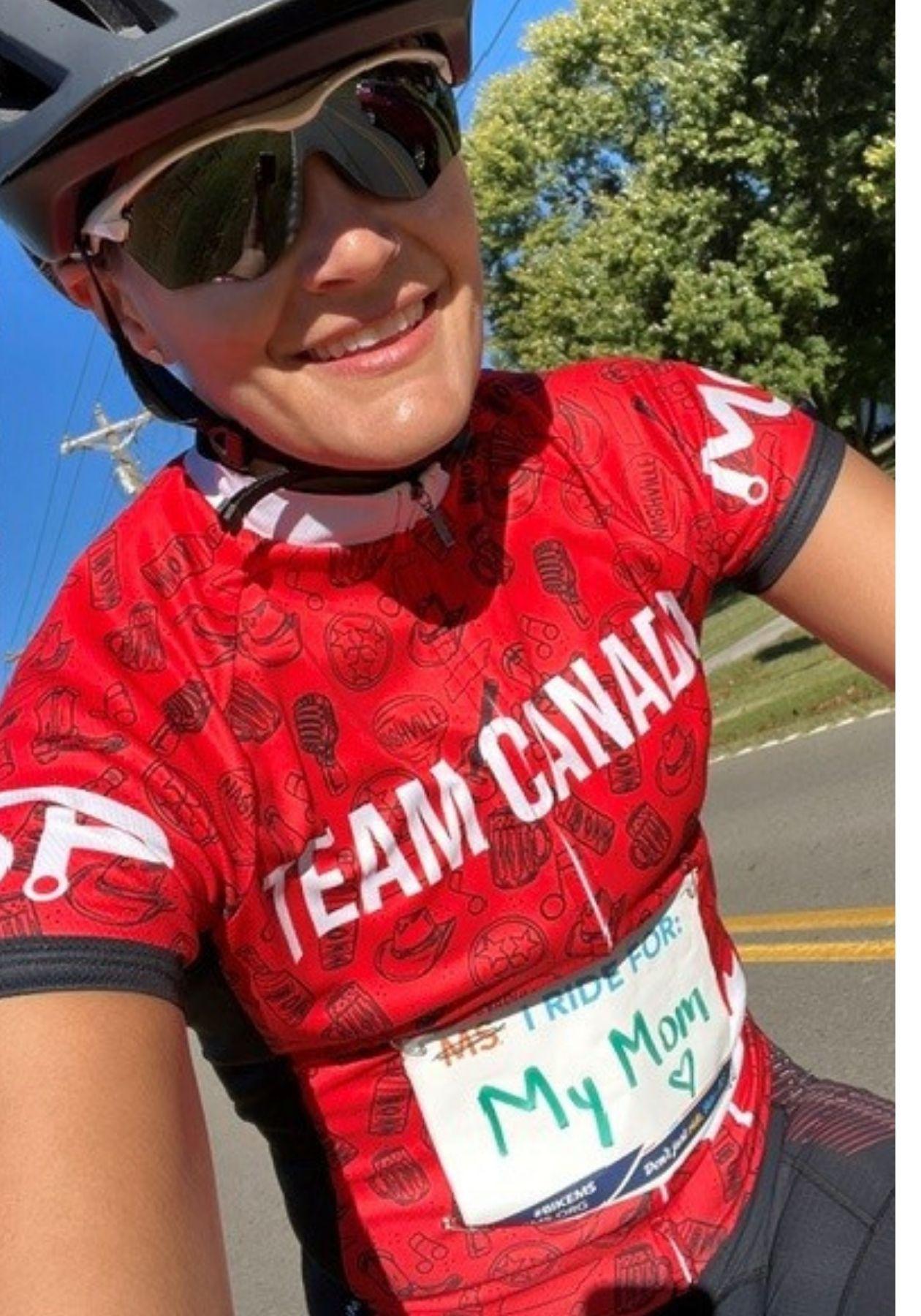 Alli smiling, on her bike, wearing a helmet, sunglasses, and red Team Canada jersey with an "I ride for mom" sign on the front.